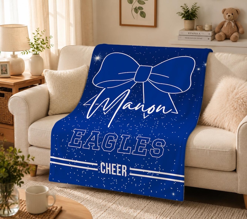 May include: A royal blue fleece blanket featuring a large white outline of a hair bow at the top, the name Manon in white script, the word EAGLES in white block letters, and the word CHEER in white text between two horizontal white lines.
