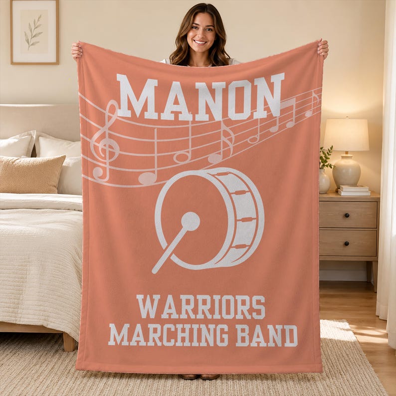 Pu&ograve; includere: Una coperta in pile color corallo con il testo bianco MANON in alto, un pentagramma musicale bianco con note al centro, una grafica bianca di una grancassa con una bacchetta e il testo bianco WARRIORS MARCHING BAND in basso.