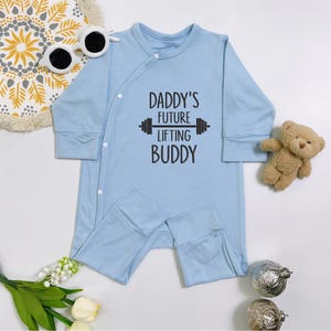 Peut inclure: Un pyjama bleu clair pour bébé avec des manches longues et des pieds attachés. Le texte "DADDY'S FUTURE LIFTING BUDDY" est imprimé en noir. Un petit ours en peluche et des lunettes de soleil sont à côté du pyjama.