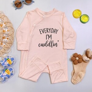 May include: A light pink baby onesie with long sleeves and attached feet. The text "EVERYDAY I'M cuddlin'" is printed in black script on the front. The onesie is surrounded by baby accessories, including socks, sunglasses, and sandals.