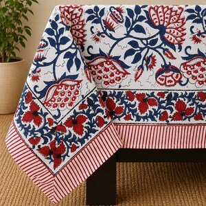 May include: A white tablecloth featuring a repeating floral pattern in deep blue and red. The design includes large stylized blossoms and leaves, bordered by a horizontal band of red and blue flowers and a final edge of red and white stripes.