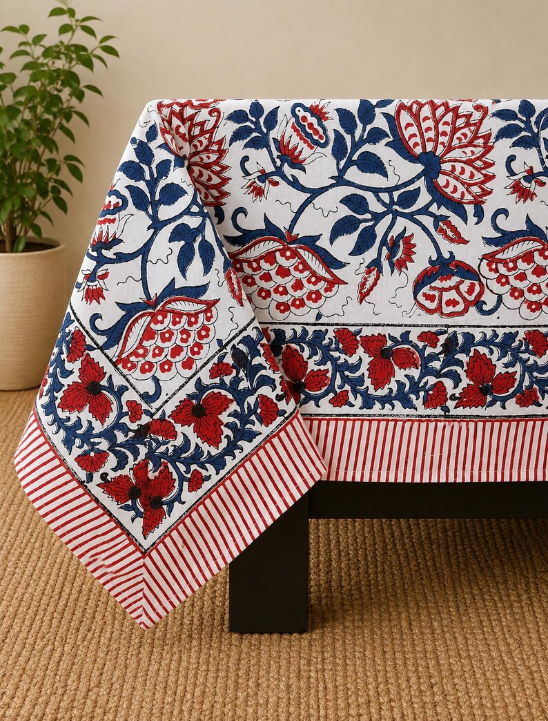 May include: A white tablecloth featuring a repeating floral pattern in deep blue and red. The design includes large stylized blossoms and leaves, bordered by a horizontal band of red and blue flowers and a final edge of red and white stripes.