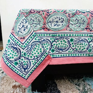 Può includere: Una tovaglia rettangolare caratterizzata da un motivo stampato a blocchi nei colori rosa, blu scuro e verde. Il design include motivi ovali contenenti forme simili ad alberi, bordi floreali e un orlo rosa tinta unita lungo i bordi.