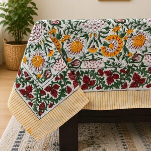 May include: A rectangular fabric tablecloth featuring a white base with a repeating floral pattern in shades of yellow, green, and deep red. The edges are finished with a distinct border of green leafy vines and a yellow and white striped trim.