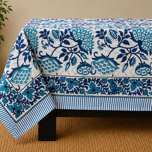 May include: A rectangular tablecloth featuring a white base with a repeating blue and teal floral pattern. The design includes large stylized flowers and leaves, a wide decorative border with smaller floral motifs, and a blue and white striped edge.