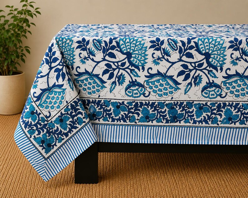 May include: A rectangular tablecloth featuring a white base with a repeating blue and teal floral pattern. The design includes large stylized flowers and leaves, a wide decorative border with smaller floral motifs, and a blue and white striped edge.