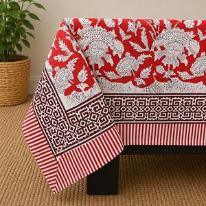May include: A rectangular tablecloth featuring a vibrant red background with a white floral print. The border displays a dark red geometric pattern and a striped edge. The fabric drapes over a dark surface, showcasing a detailed block-print style design.