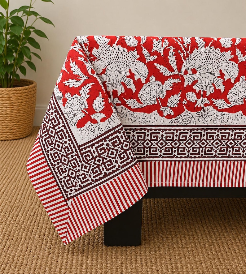 May include: A rectangular tablecloth featuring a vibrant red background with a white floral print. The border displays a dark red geometric pattern and a striped edge. The fabric drapes over a dark surface, showcasing a detailed block-print style design.