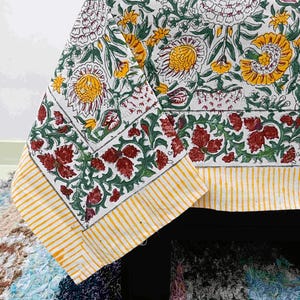 Könnte beinhalten: Eine weiße Tischdecke mit einem Blumenmuster mit gelben, roten und grünen Blumen und Blättern. Der Rand hat ein gelb-weiß gestreiftes Design. Die Tischdecke ist über einer schwarzen Oberfläche drapiert.
