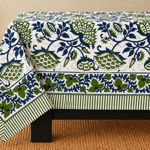May include: A rectangular tablecloth featuring a white background with a repeating pattern of large dark blue and olive green floral motifs. The border includes a decorative band of green leaves and blue vines, finished with a striped green edge.