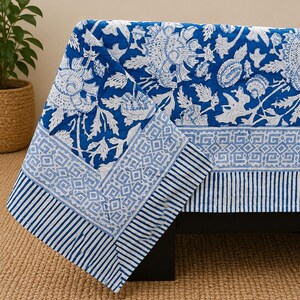 May include: A rectangular fabric tablecloth featuring a vibrant blue and white floral pattern. The design includes a central section with large stylized flowers, bordered by a geometric diamond motif and a striped edge in alternating blue and white.
