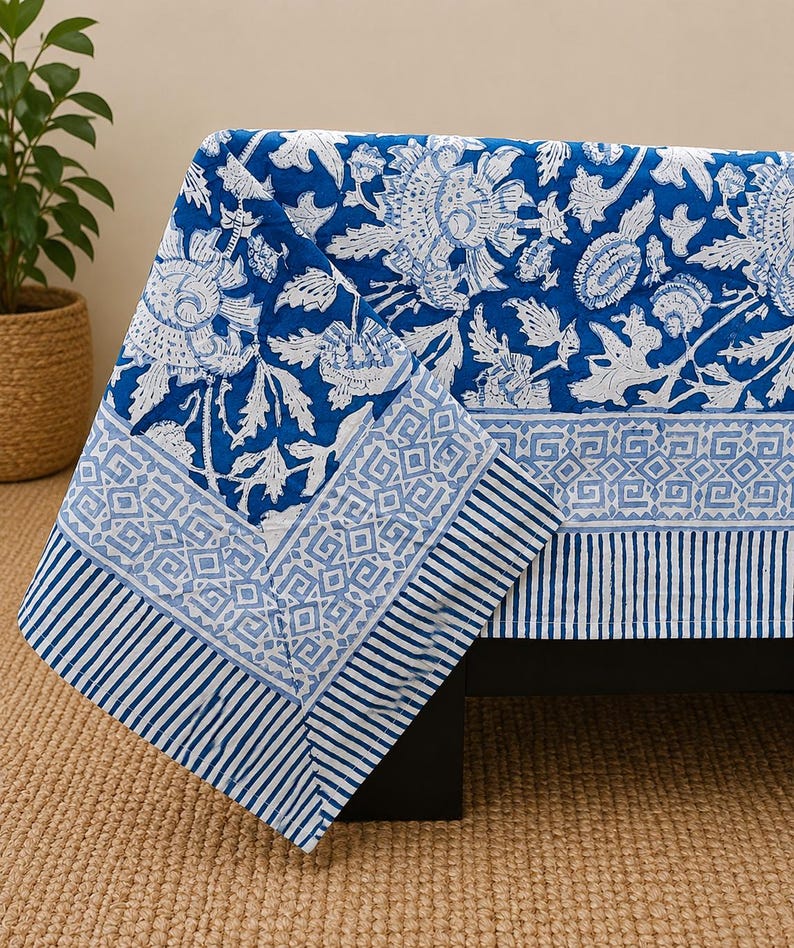 May include: A rectangular fabric tablecloth featuring a vibrant blue and white floral pattern. The design includes a central section with large stylized flowers, bordered by a geometric diamond motif and a striped edge in alternating blue and white.