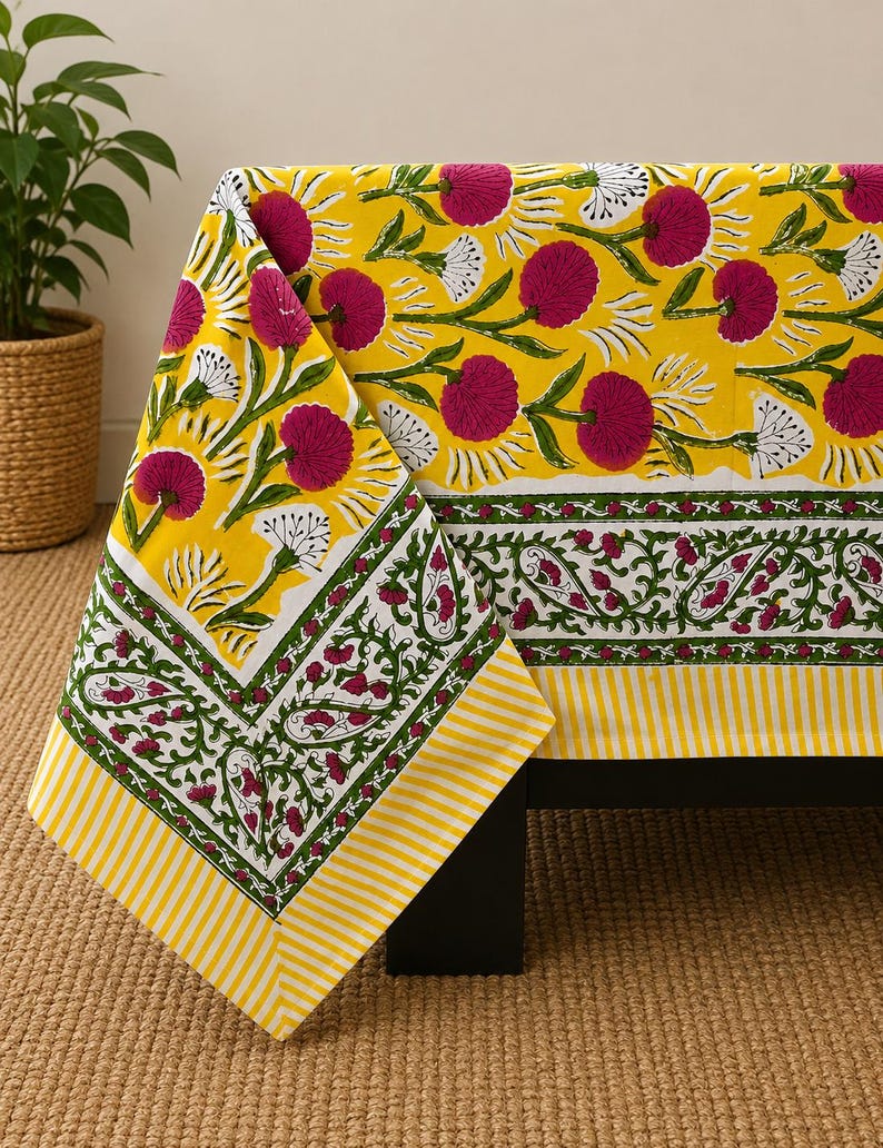 May include: A rectangular tablecloth featuring a bright yellow background with a repeating pattern of magenta flowers and white accents. The border displays a green vine motif with magenta floral details and a yellow and white striped edge.