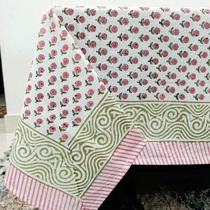 Könnte beinhalten: Weißes Tischtuch mit einem sich wiederholenden Muster aus rosa Blütenclustern und grünen Blättern. Der Rand hat ein grünes Wirbeldesign und rosa und weiße Streifen. Das Tischtuch ist über eine dunkle Oberfläche gefaltet.