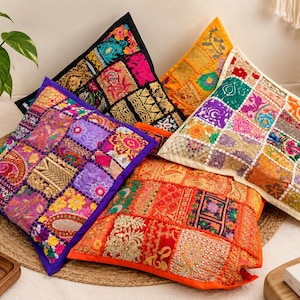 Könnte beinhalten: Eine Sammlung von vier quadratischen Dekokissen mit komplexen Patchwork-Designs, lebendigen, mehrfarbigen Stickereien, Pailletten und metallischen Fadenakzenten. Jedes Kissen zeigt eine einzigartige Anordnung von Stoffquadraten in den Farben Violett, Orange, Schwarz und Creme.