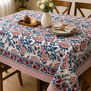 May include: A rectangular tablecloth featuring a white base with a repeating floral pattern in shades of deep blue and red. The design includes a wide border with a striped edge, covering a wooden table set with a vase, plates, and a candle.