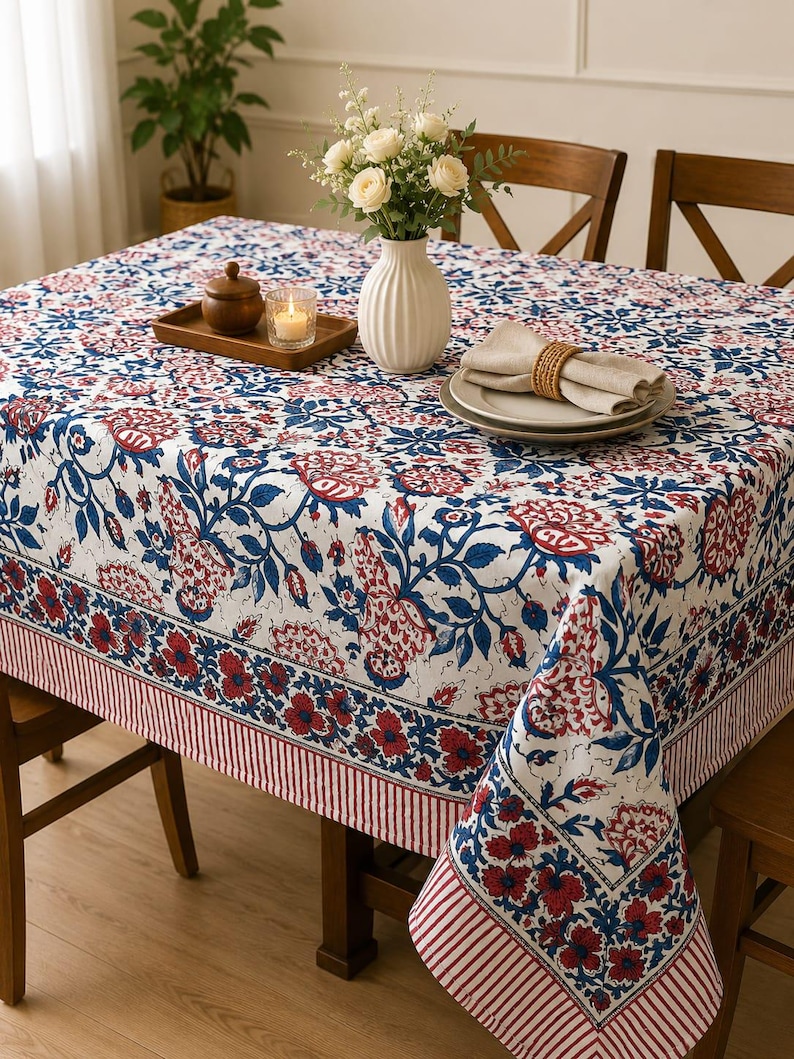 May include: A rectangular tablecloth featuring a white base with a repeating floral pattern in shades of deep blue and red. The design includes a wide border with a striped edge, covering a wooden table set with a vase, plates, and a candle.