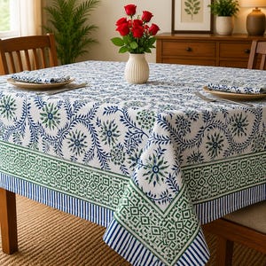 May include: A rectangular tablecloth featuring a white base with a blue and green floral vine pattern, a green geometric border, and a blue and white striped edge. The fabric drapes over a wooden table set with plates, napkins, and a vase of red roses.