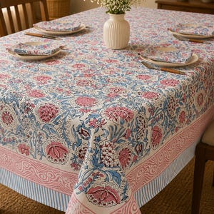 May include: A rectangular tablecloth featuring a floral pattern in shades of pink and blue on a white background, with a decorative border of pink wavy lines and blue stripes. The fabric covers a table set with matching napkins and a white vase.