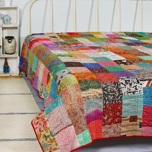 Könnte beinhalten: Eine farbenfrohe Patchwork-Steppdecke mit rotem Rand bedeckt ein Bett. Die Steppdecke besteht aus verschiedenen Stoffquadraten in Rot-, Blau-, Grün-, Gelb- und Brauntönen. Die Steppdecke hat ein genähtes Design.