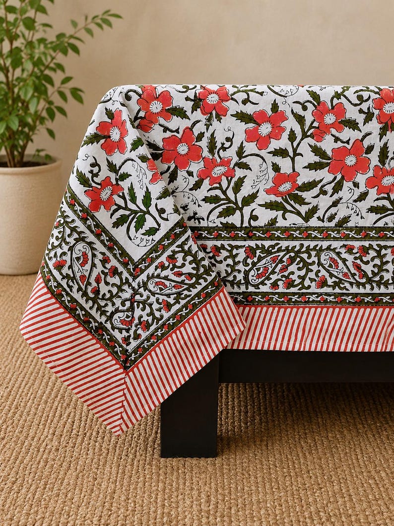 May include: A white tablecloth featuring a repeating pattern of red flowers and green vines. The border includes a decorative band of green paisley motifs and a final edge of red and white diagonal stripes, creating a structured visual design.