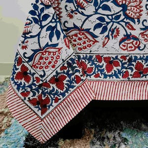 May include: A white tablecloth featuring a repeating floral pattern in red and blue. The design includes large stylised blossoms and leafy vines, bordered by a horizontal band of smaller red flowers and a final edge of thin red and white stripes.