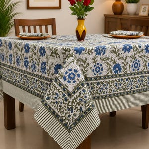 May include: A square tablecloth featuring a white base with a repeating blue floral and green vine pattern. The border includes a decorative band of blue flowers and green leaves, finished with a striped edge in white and dark green tones.