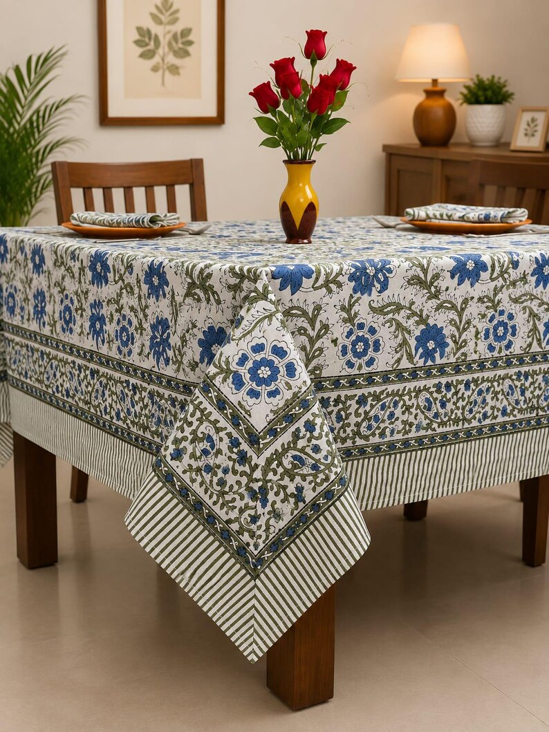 May include: A square tablecloth featuring a white base with a repeating blue floral and green vine pattern. The border includes a decorative band of blue flowers and green leaves, finished with a striped edge in white and dark green tones.
