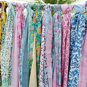 Könnte beinhalten: Eine Sammlung farbenfroher, gemusterter Schals, die an einer Stange hängen. Die Schals zeigen verschiedene Blumen- und geometrische Designs in Blau-, Rosa-, Gelb- und Grüntönen. Der Stoff wirkt leicht und für wärmeres Wetter geeignet.