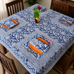 May include: A square tablecloth and matching napkins featuring a blue and white floral and geometric block print pattern. The table setting includes orange plates, silver forks, and silver spoons arranged on the fabric, with a red rose in a vase.