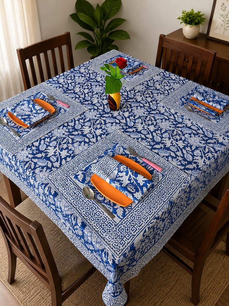 May include: A square tablecloth and matching napkins featuring a blue and white floral and geometric block print pattern. The table setting includes orange plates, silver forks, and silver spoons arranged on the fabric, with a red rose in a vase.