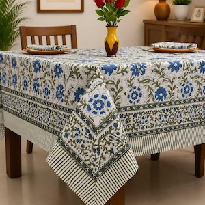 Könnte beinhalten: Eine quadratische Tischdecke mit weißem Untergrund, einem wiederkehrenden blauen Blumenmuster und grünen Rankenmotiven. Der Rand besteht aus einem dekorativen Band mit blauen Blumen und einem breiten Saum mit dünnen, vertikalen Streifen.
