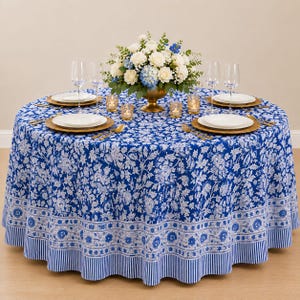 Peut inclure: Une nappe ronde ornée d'un motif floral bleu et blanc avec une bordure rayée. La table est dressée avec des assiettes blanches, des verres à vin transparents, des couverts dorés, des bougeoirs en verre et un centre de table floral.