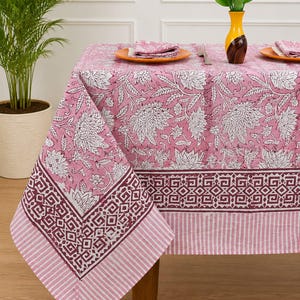 May include: A rectangular tablecloth featuring a pink and white floral pattern with a dark maroon geometric border and a striped edge. The fabric displays large white flower motifs on a pink background, finished with a distinct patterned trim.