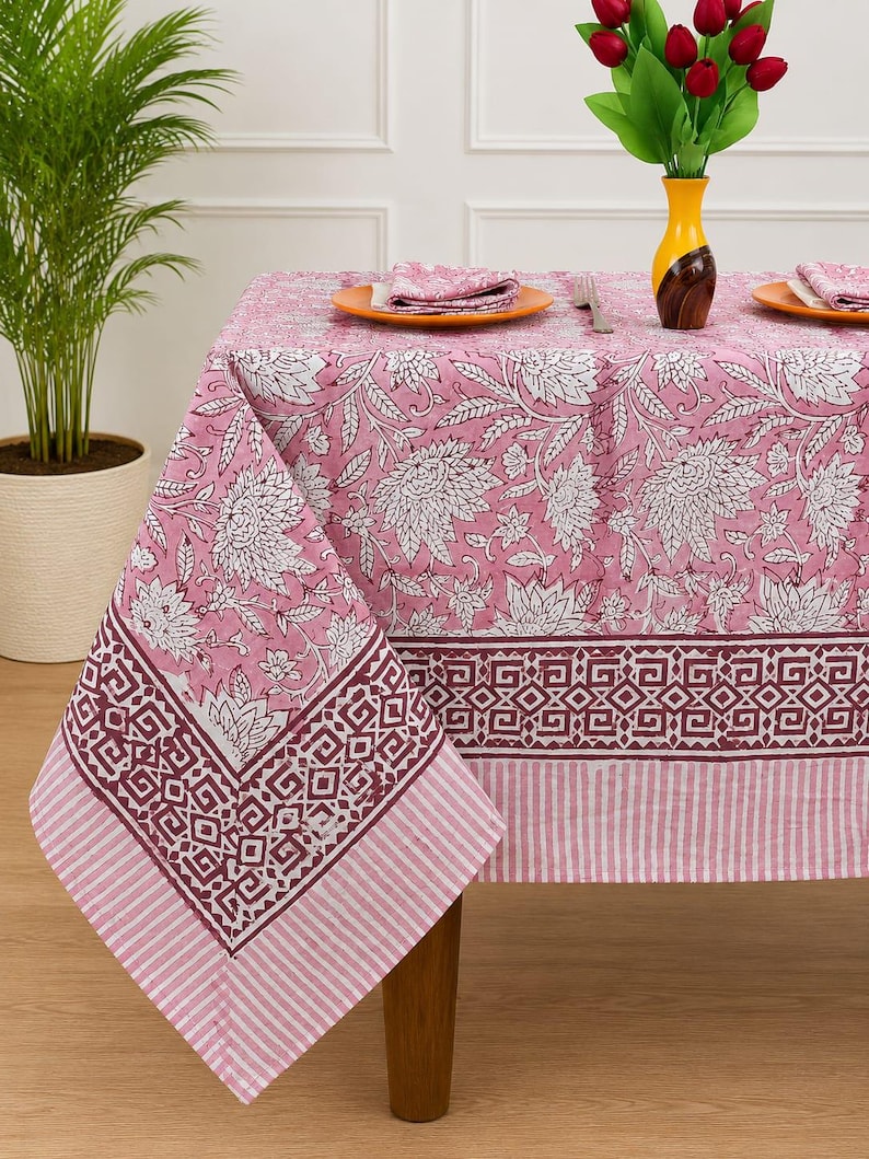 May include: A rectangular tablecloth featuring a pink and white floral pattern with a dark maroon geometric border and a striped edge. The fabric displays large white flower motifs on a pink background, finished with a distinct patterned trim.