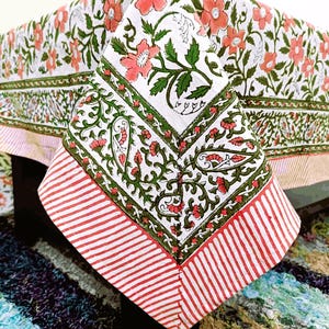 Könnte beinhalten: Ein weißes Tischtuch mit einem Blumenmuster aus rosa Blüten und grünen Blättern. Der Rand hat ein grünes und rotes Design, und der Rand hat rot-weiße Streifen. Das Tischtuch ist aus einem weichen, leichten Stoff gefertigt.