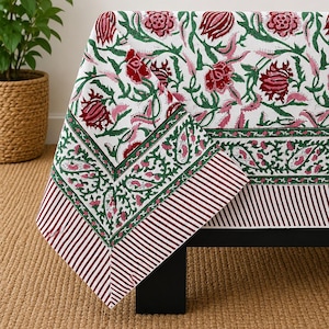 Könnte beinhalten: Eine weiße Tischdecke mit einem sich wiederholenden Blumenmuster in Dunkelrot und Grün. Das Design zeigt große Blüten und Blattreben, umrandet von einem dekorativen Band aus grünem Blattwerk und einem Abschluss aus dünnen, parallelen Streifen.
