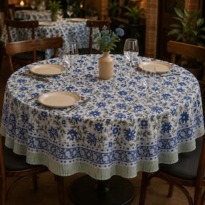 May include: A circular tablecloth featuring a blue floral design on a white backdrop. The table is set with plates, cutlery, and wine glasses. A vase of blue flowers is in the centre.