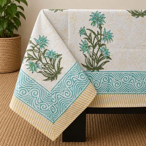 May include: A white tablecloth featuring a block-printed pattern of teal and green floral motifs. The border displays a teal wave design followed by a yellow and white striped edge, with a faint, light-colored botanical print across the fabric.
