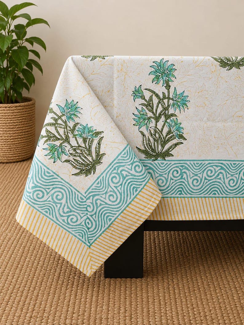 May include: A white tablecloth featuring a block-printed pattern of teal and green floral motifs. The border displays a teal wave design followed by a yellow and white striped edge, with a faint, light-colored botanical print across the fabric.