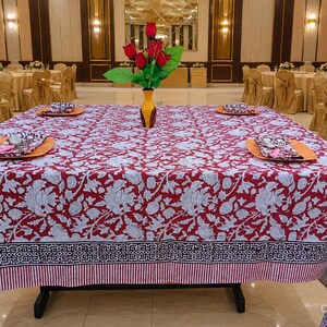Könnte beinhalten: Ein quadratischer Esstisch mit einer bordeauxroten und weißen Tischdecke mit Blumenmuster und geometrischer Bordüre. In der Mitte steht eine gelbe Vase mit künstlichen roten Rosen, flankiert von orangefarbenen Tellern mit gemusterten Servietten.