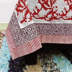 Könnte beinhalten: Eine rot-weiß gemusterte Tischdecke mit Blumenmuster. Die Tischdecke hat ein großes Blumendesign in Weiß auf rotem Hintergrund. Der Rand hat geometrische Muster und vertikale Streifen in Rot und Weiß.