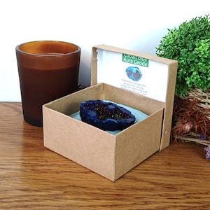 May include: A cobalt blue aura geode in a small cardboard box, with a printed label. A brown candle and a small green plant in a woven basket are in the background. The geode is a deep blue color with sparkling crystals.