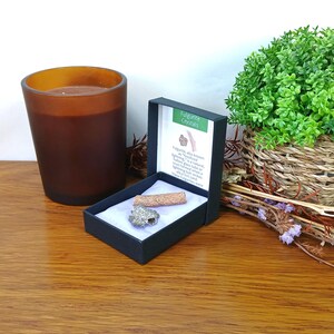 May include: A black jewellery box containing a cylindrical tan fulgurite specimen and a small, textured, metallic-coloured stone. The box lid features a white label with green text reading Fulgurite Crystals and a descriptive paragraph about the item.