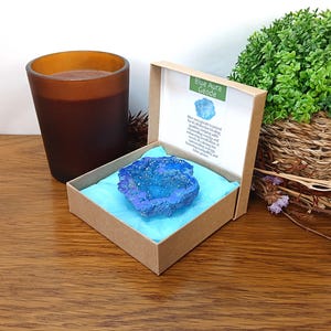 May include: A blue aura geode rests inside a square brown cardboard box lined with light blue tissue paper. The box lid features a label with the text Blue Aura Geode and a description of the stone. The geode has a crystalline, textured surface.