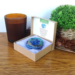 May include: A sapphire aura geode in a cardboard box with a white insert. The geode has a blue crystal center and a blue and gray outer edge. A brown candle and a small green plant in a woven basket are also in the image.