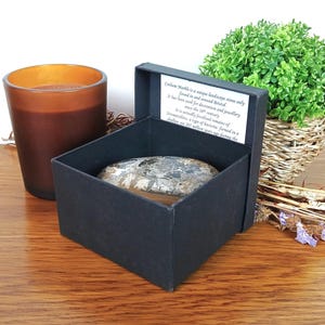 Peut inclure: Un bougeoir en verre brun foncé et une boîte noire contenant une pierre grise et marron. La boîte est ouverte, révélant la pierre à l'intérieur. Une petite plante verte dans un panier tressé est en arrière-plan.