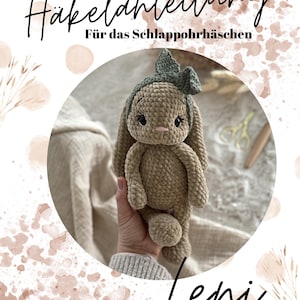 Op de afbeelding: Een gehaakte konijnenpop in beige met een groene strik en haarband. De afbeelding bevat de tekst "Häkelanleitung Für das Schlappohrhaschen" en de naam "Leni". De pop wordt in een hand gehouden.