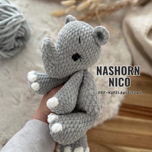 Könnte beinhalten: Ein graues, gehäkeltes Nashorn-Stofftier mit weißen Akzenten an den Füßen, schwarzen gestickten Augen und Ohren. Der Text NASHORN NICO PDF-HÄKELANLEITUNG ist rechts im Bild zu sehen und weist auf eine Häkelanleitung hin.