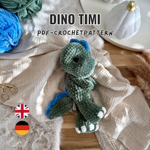 May include: A green plush dinosaur toy with blue spikes along its back and white accents on its feet. The text DINO TIMI PDF-CROCHETPATTERN is displayed above the toy. The dinosaur has large dark eyes and a textured, soft yarn construction.